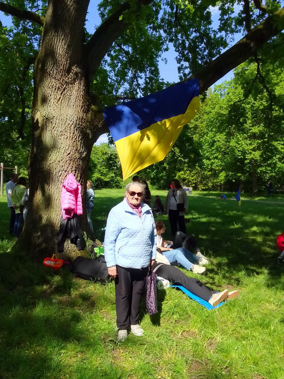 Eine ältere Frau mit Sonnenbrille steht vor einem Baum auf einer Wiese und schaut in die Kamera. Im Baum hängt eine Ukraine-Flagge.