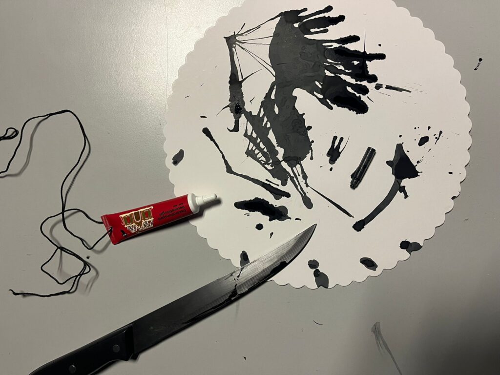 Ein scharfes Messer und eine Farbtube auf einem Pappteller . Der Pappteller ist mit schwarzer Farbe verschmiert.