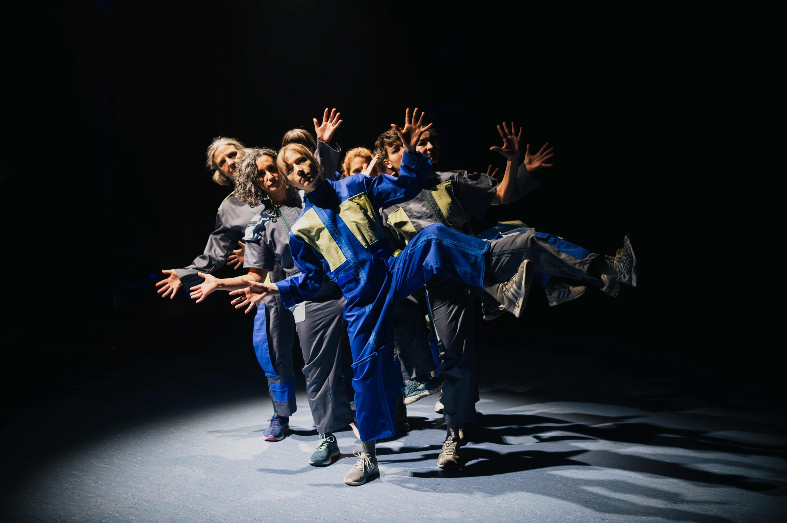 Seven women in blue jumpsuits are standing on one leg, spreading theirs fingers.