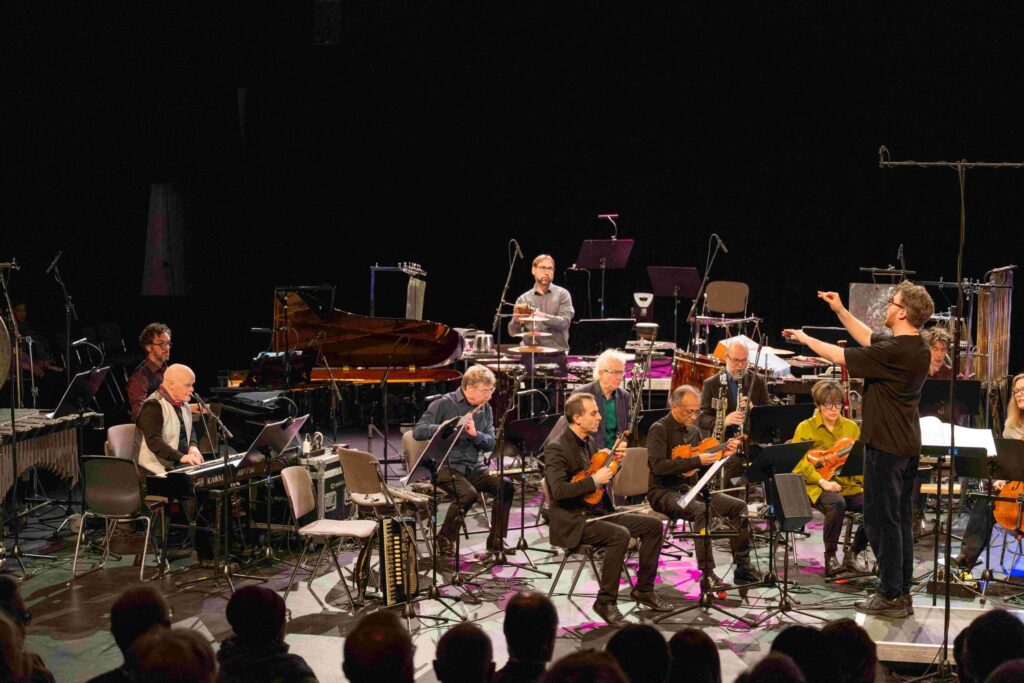 Musiker*innen des Ensemble Modern. Rechts steht der Dirigent mit erhobenen Armen, im Vordergrund Köpfe von Zuschauer*innen.