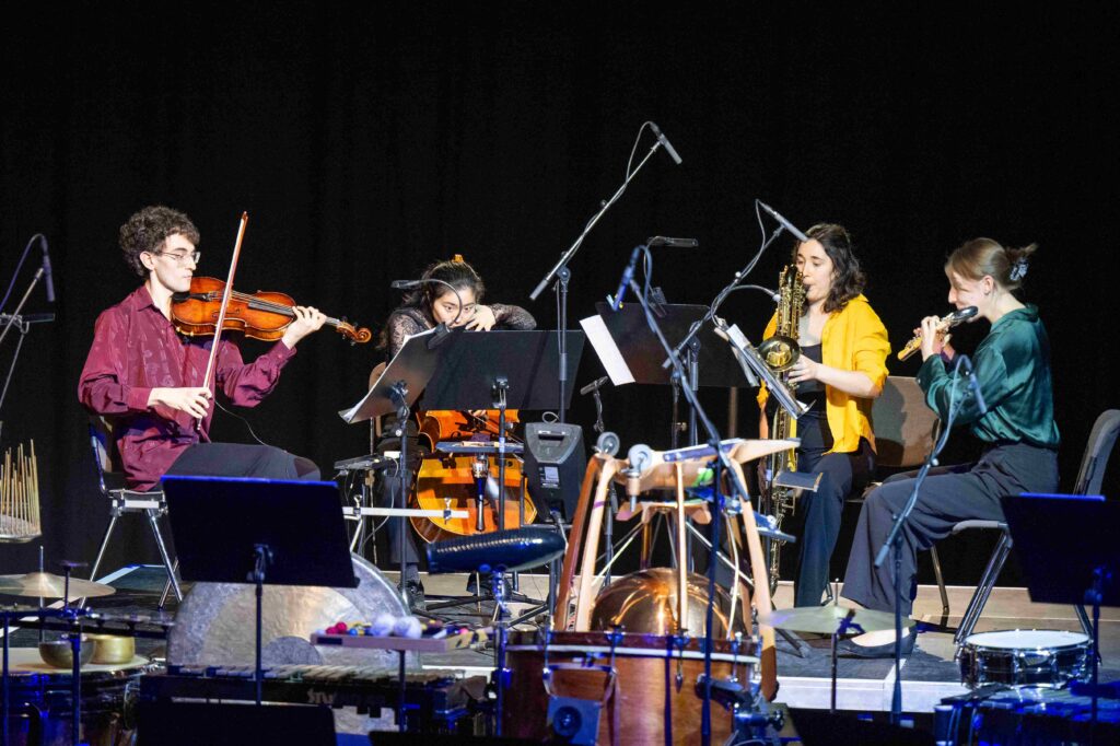 Vier junge Musiker*innen spielen Violine, Cello, Saxophone und Querflöte, umgeben von Mikrophonen und Notenständern.