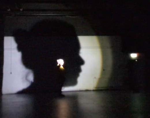 Der Kopf einer Person wird hell angeleuchtet. Im Hintergrund erscheint der Schatten groß an der Wand.