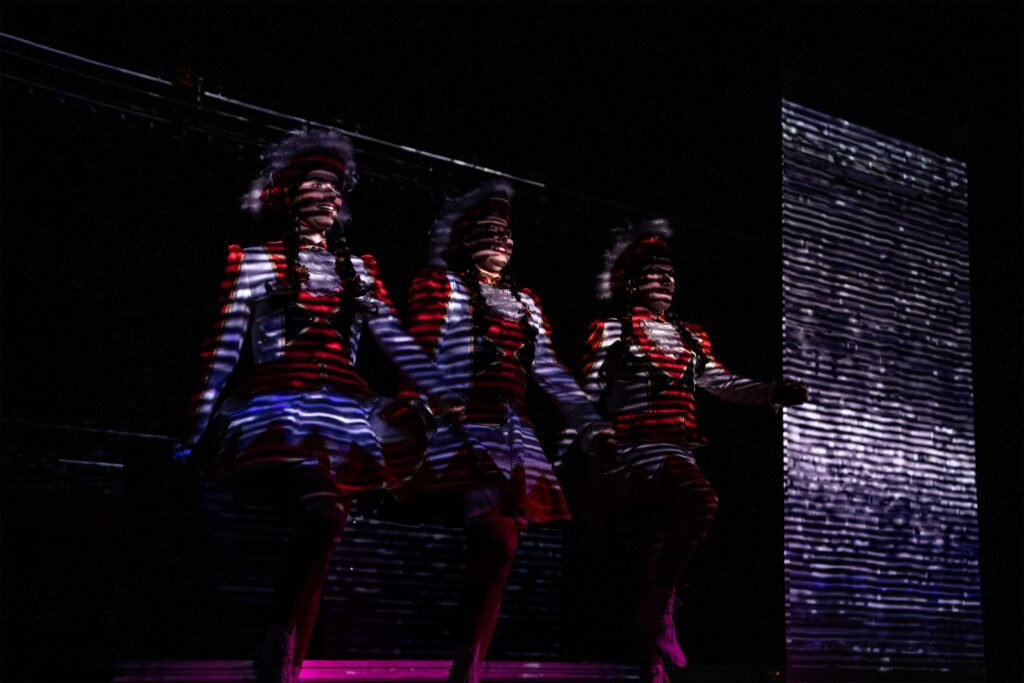 Drei Performer*innen in rot-weißer Gardeuniform stehen im gestreiften Licht einer Videoprojektion.
