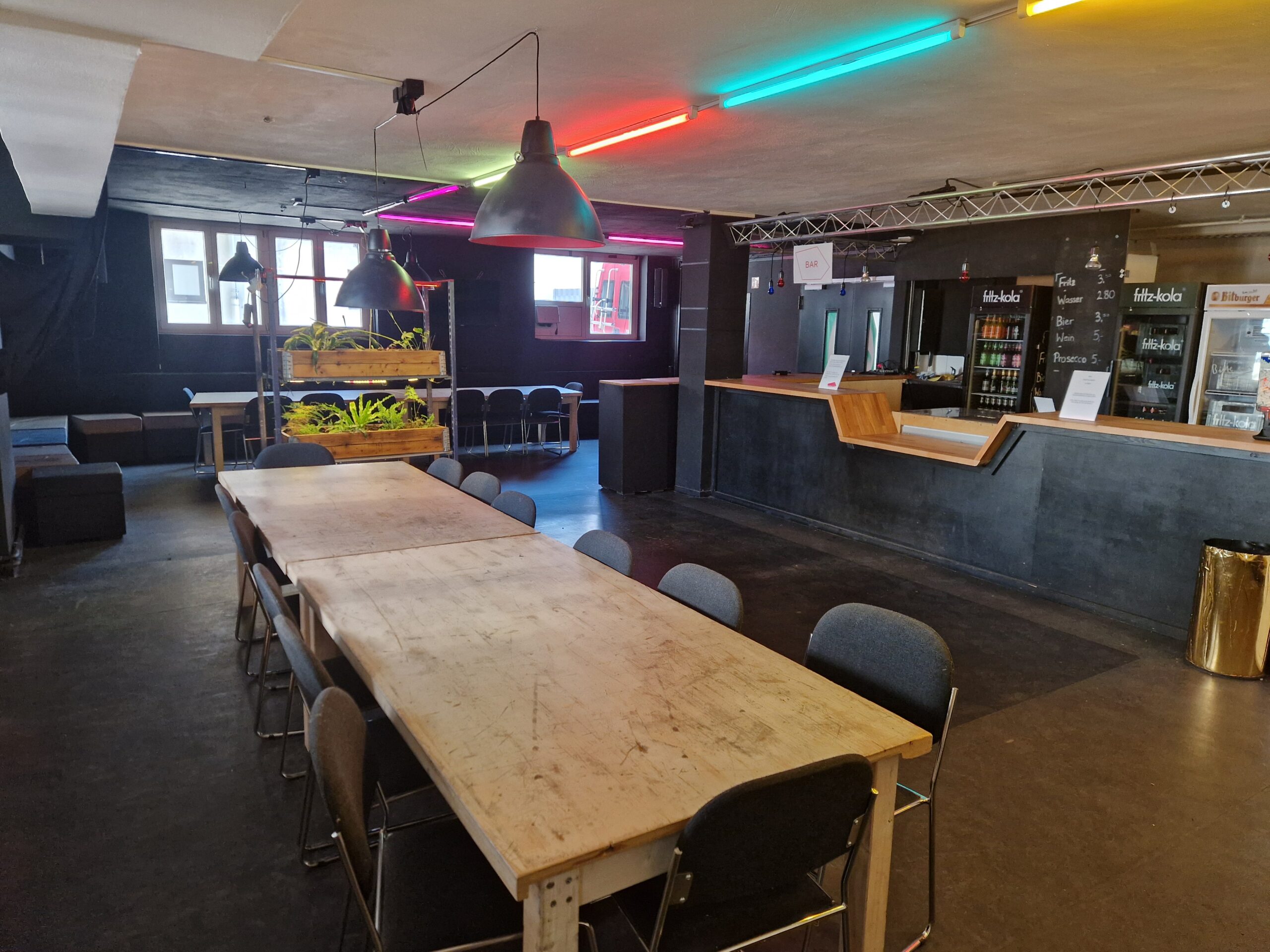 Das leere Foyer des Frankfurt LAB. Große alte Holztische , eine Bar und ein Gestell mit Pflanzen. Der Raum ist schwarz gestrichen und von bunten Neonröhren beleuchtet.