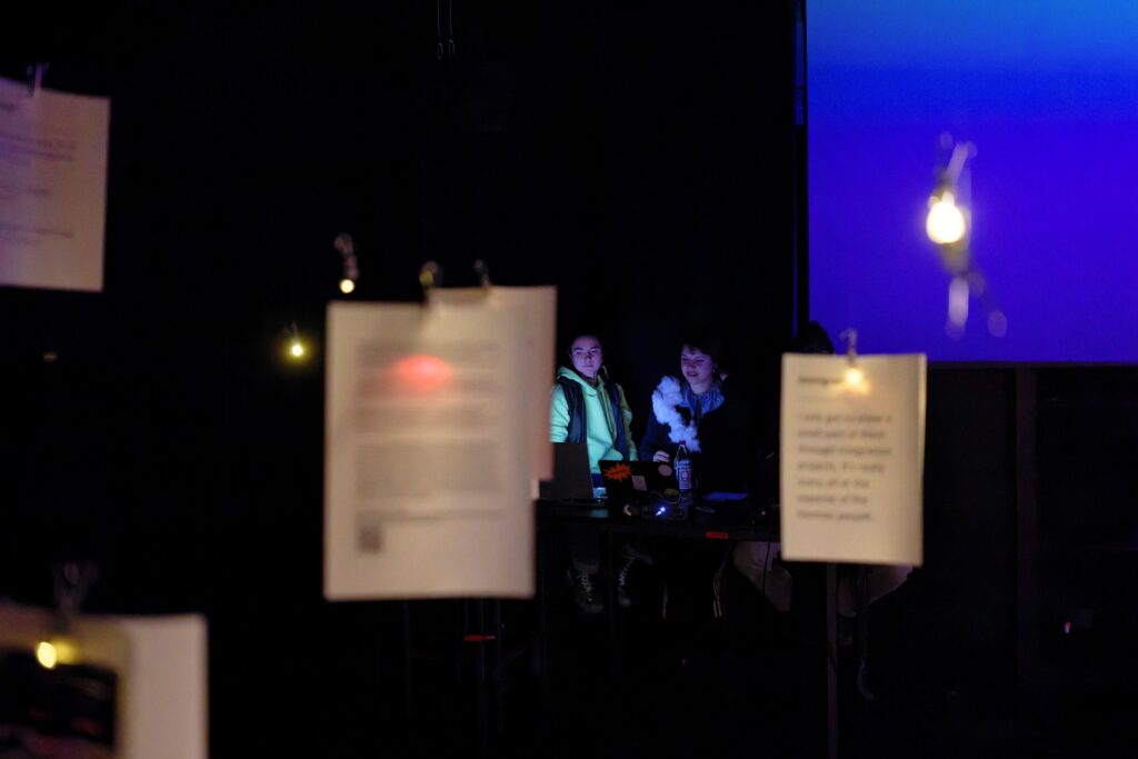 Im Vordergrund hängen bedruckte Zettel. Im Hintergrund stehen zwei Performerinnen vor Laptops.