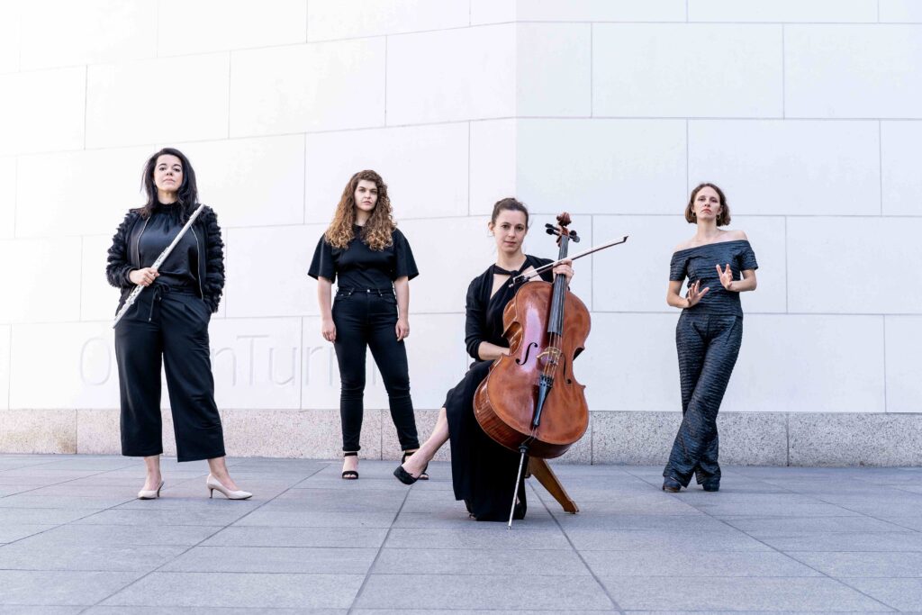Vier schwarz gekleidete Frauen vor einer weißen Wand. Eine hält eine Querflöte in der Hand und eine ein Cello.