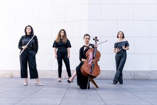 Vier schwarz gekleidete Frauen vor einer weißen Wand. Eine hält eine Querflöte in der Hand und eine ein Cello.