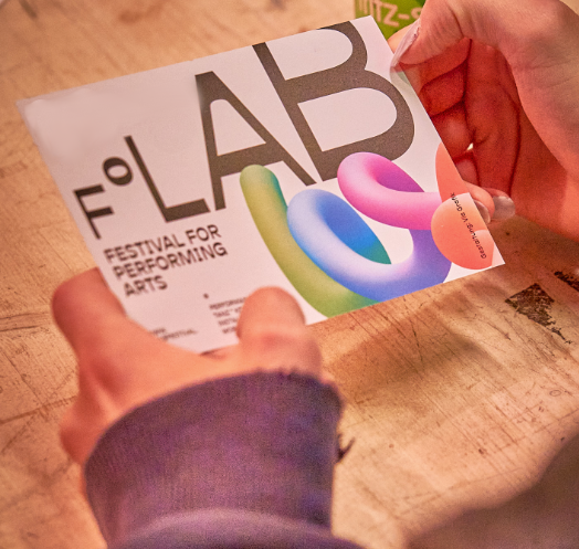 Eine Person hält eine Postkarte des F°LAB Festivals in der Hand auf der bunte Schlangenlinien zu sehen sind.