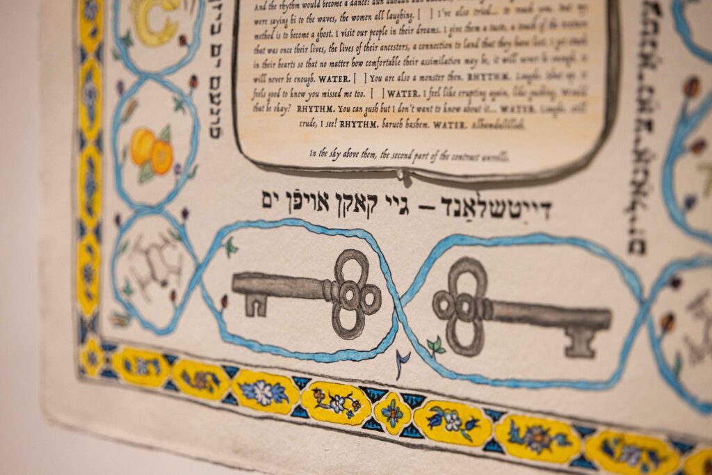 Das Foto zeigt einen Ausschnitt eines Posters, auf dem hebräischer und englischer Text abgedruckt sind, umgeben von einer gezeichneten Bordüre in blau und geld mit Blumen und zwei großen, antiken Schlüsseln.