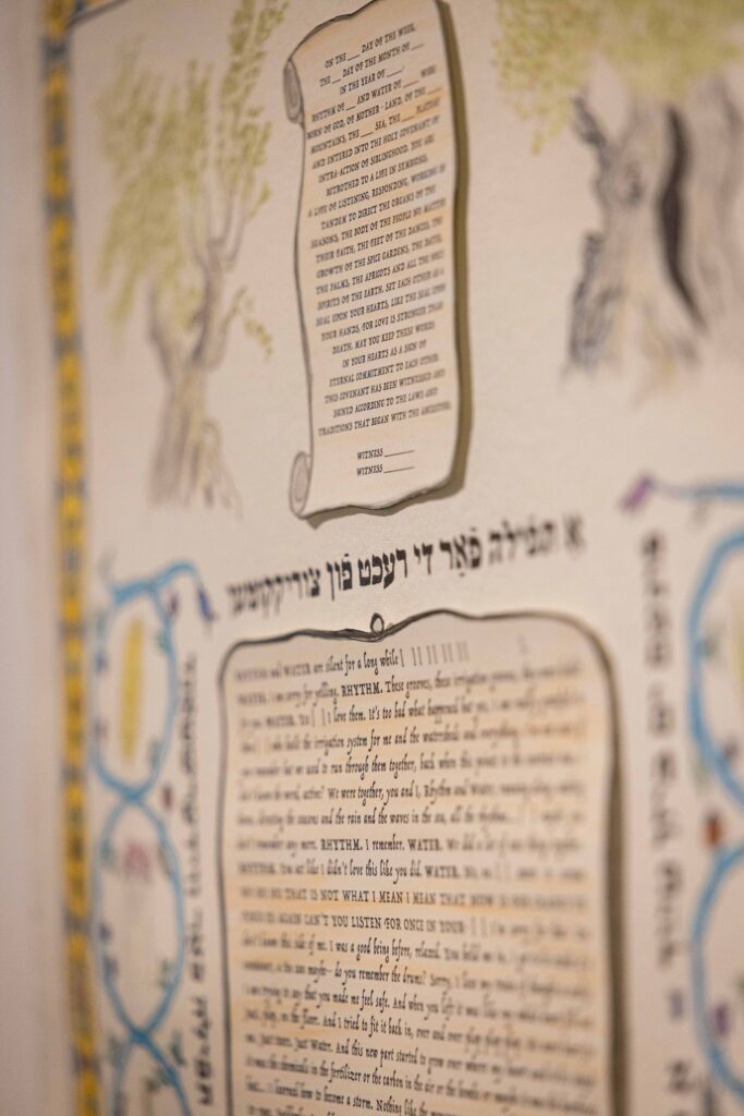 Das Foto zeigt einen Ausschnitt eines Posters, auf dem hebräischer und englischer Text im Layout von Schriftrollen abgedruckt sind, umgeben von einer gezeichneten Bordüre in blau und gelb sowie zwei gezeichneten Pflanzen.