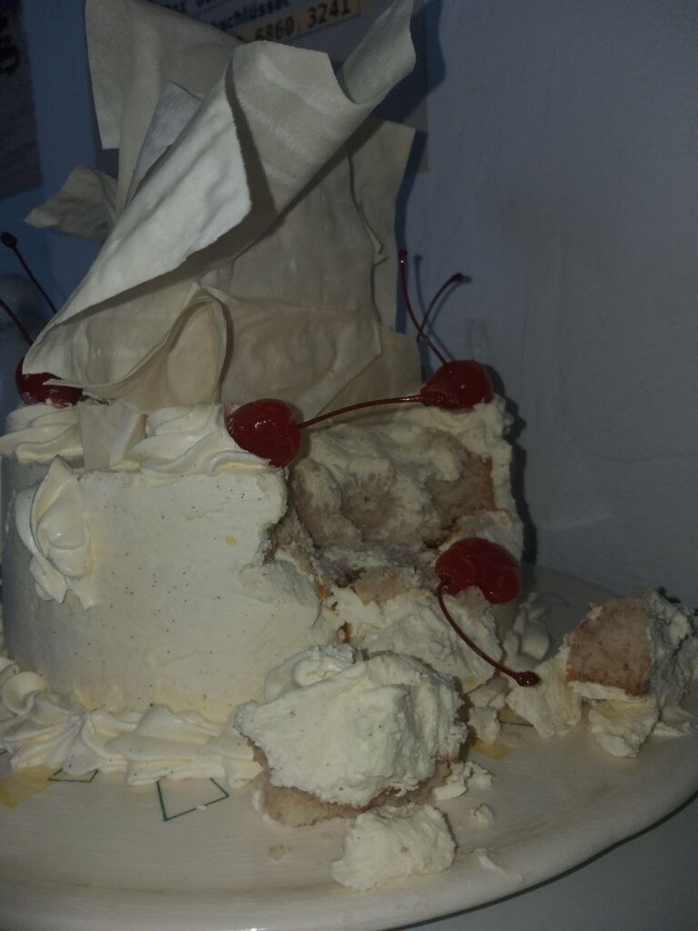 Das Foto zeigt eine angebissene, runde weiße Torte mit Sahne und großem Topping sowie einigen Cocktailkirschen.