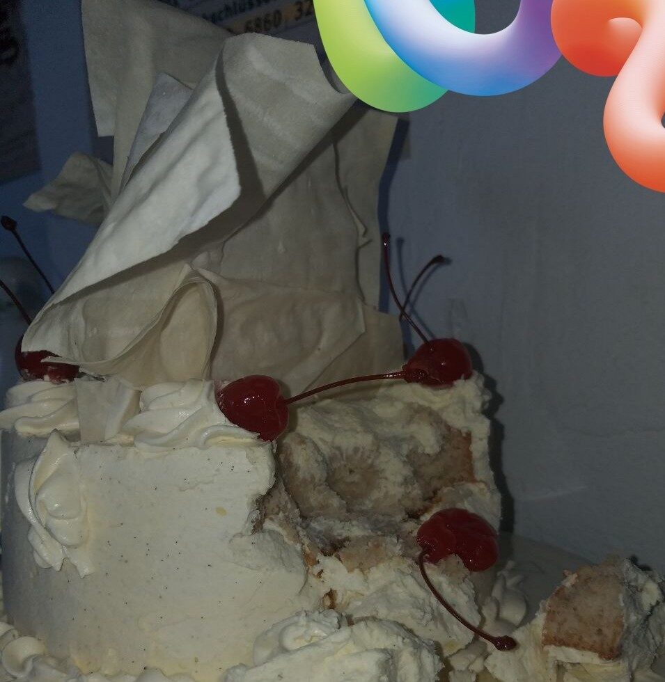 Das Foto zeigt eine angebissene, runde weiße Torte mit Sahne und großem Topping sowie einigen Cocktailkirschen.