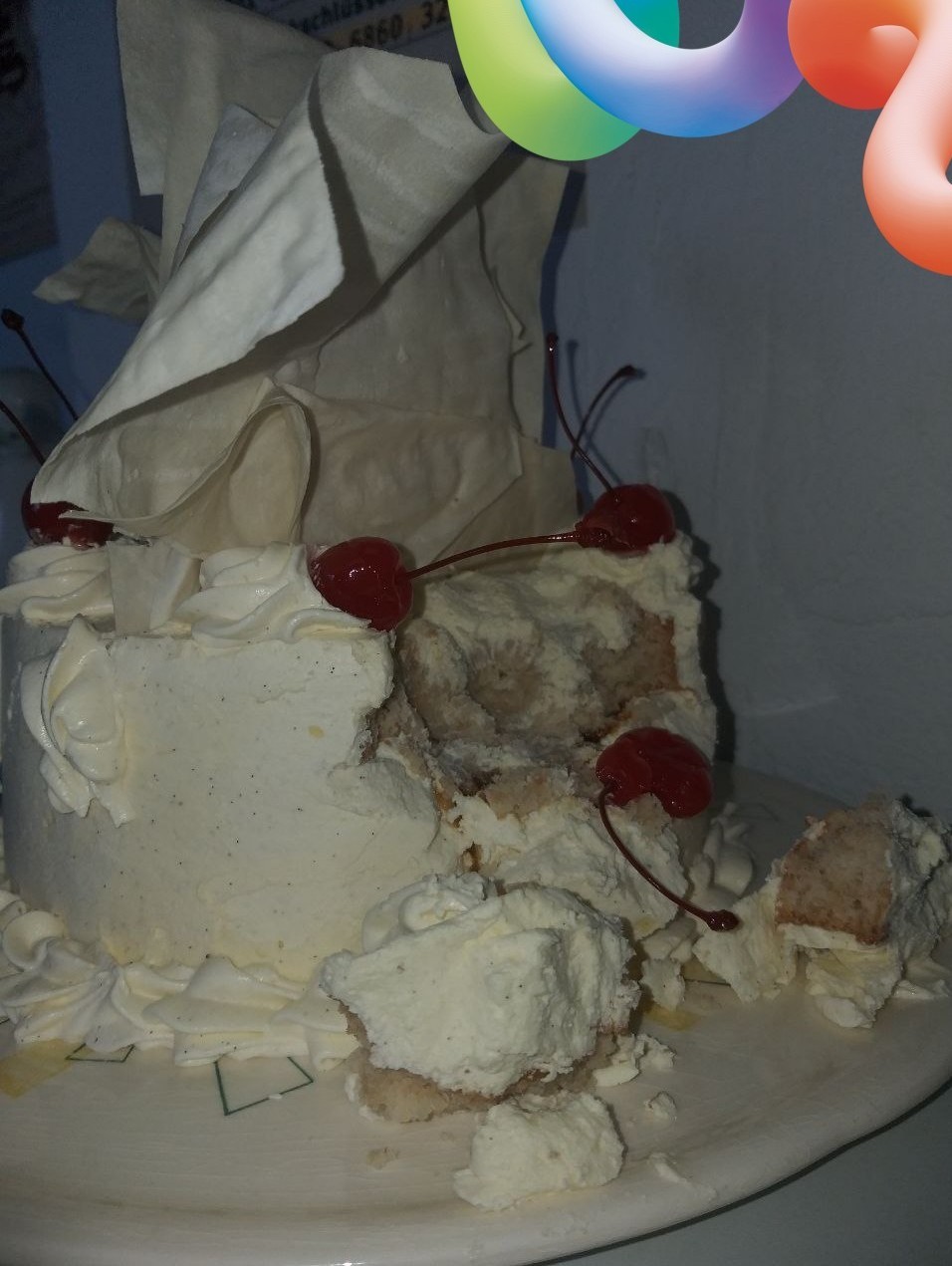 Das Foto zeigt eine angebissene, runde weiße Torte mit Sahne und großem Topping sowie einigen Cocktailkirschen.