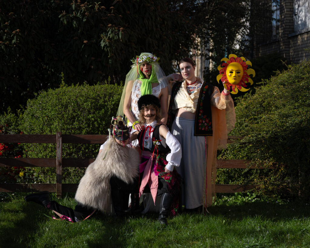 Das Bild zeigt eine Gruppe von vier Performer*innen in einem Garten, die aufwändig kostümiert sind. Eine Person ist als Mann verkleidet, eine weitere als Braut, eine dritte trägt Fellmantel und Tiermaske und die vierte Person hält neben den Kopf die Maske einer Sonne.
