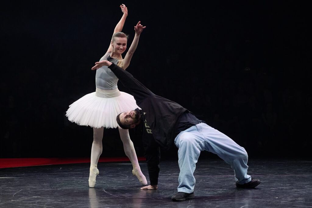 Ein Tänzer im Hiphopkostüm macht eine Figur auf dem Boden, während hinter ihm eine Ballerina posiert und auf ihn hinab lächelt.