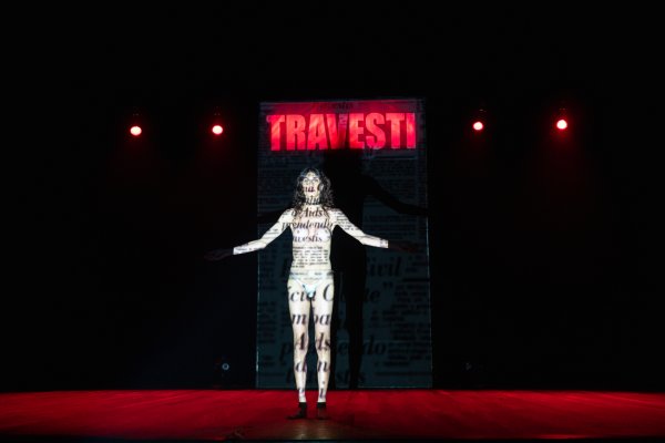 Eine Person in Unterwäsche steht mit ausgebreiteten Armen unter einer roten Leuchtschrift "Travesti". Auf ihren Körper ist Text projiziert.