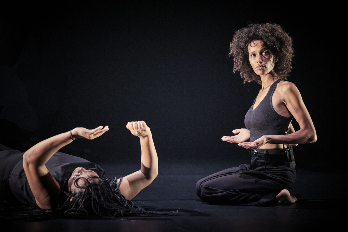 Two black women in black pants and shirts. One is lying down, lifting her hands. One is kneeling with her hands held out in front of her.