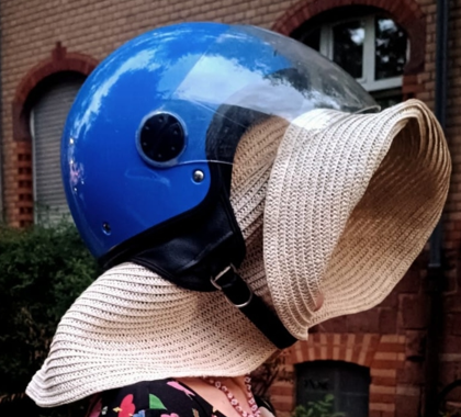 Der Kopf einer Frau in Nahaufnahme, die einen blauen Motorradhelm trägt und darunter einen Stohhut, der ihr Gesicht verdeckt.