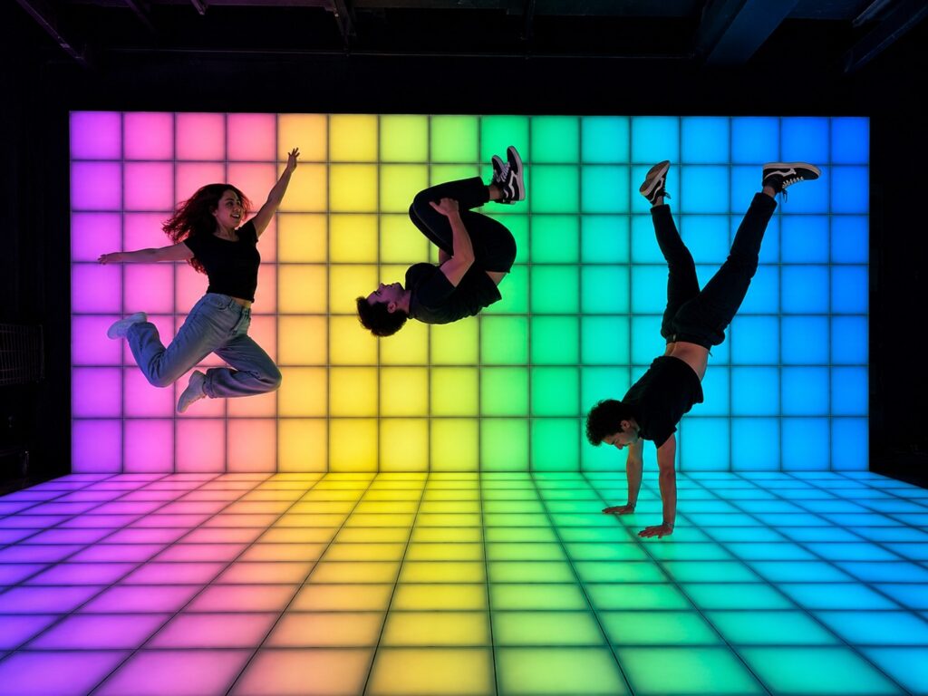 Ein mit bunten Pixel ausgefüllter Raum. In ihm springen drei junge Menschen akrobatisch in die Luft.