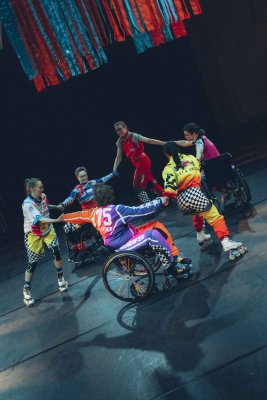 Sechs bunt bekleidete Performer*innen bilden einen Kreis und halten sich and den Händen. Drei sitzen im Rollstuhl, die anderen sind auf Inline-Skates.